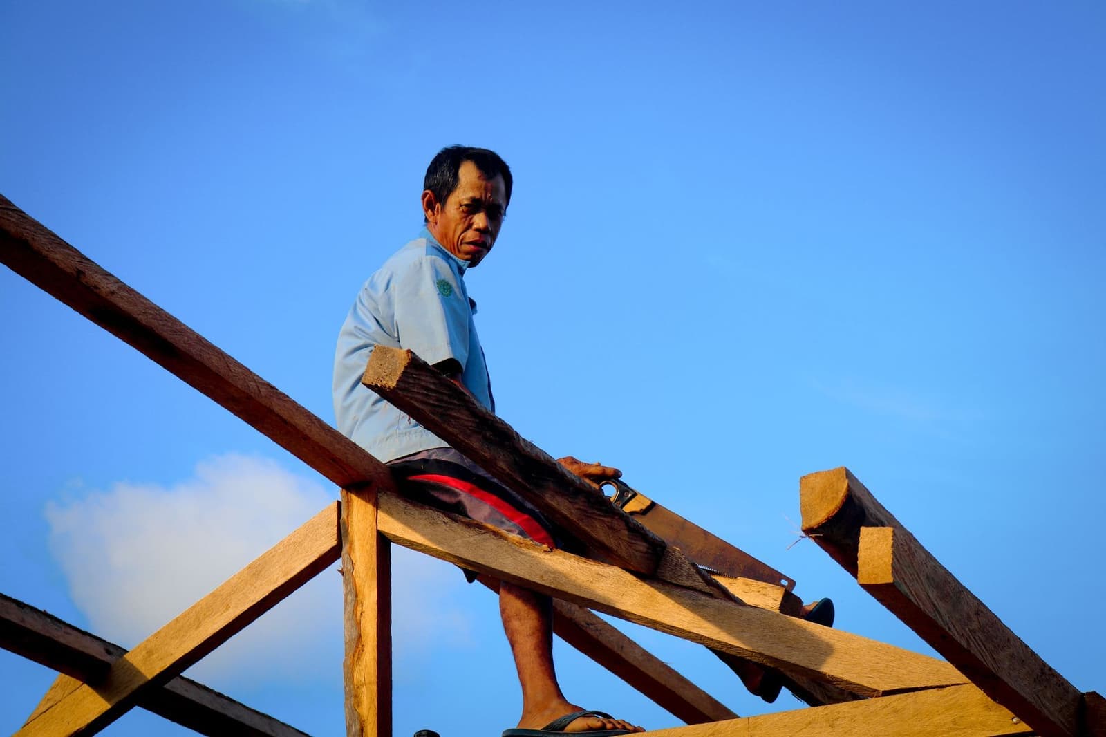 Worker sitting on timber roof rafters against blue sky
